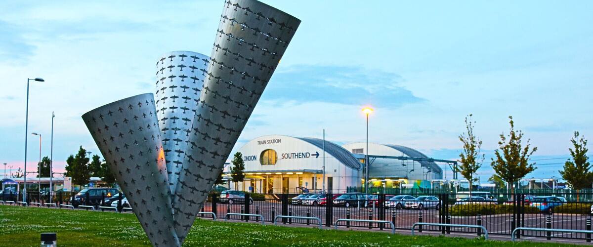 A modern sculpture in a public square, with a train station and cars in the background.