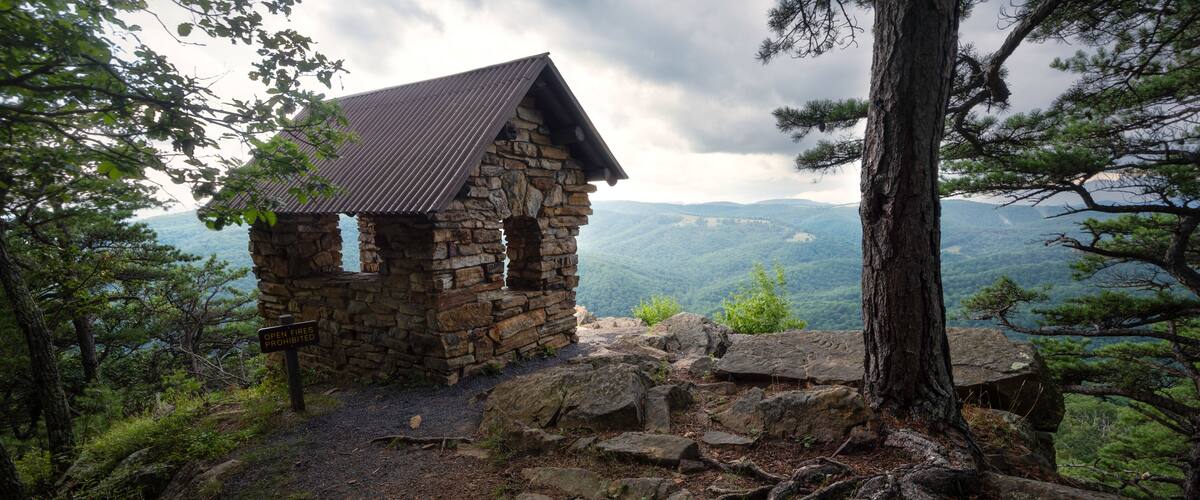 A stone cabin with a metal roof surrounded by trees and rocky terrain.