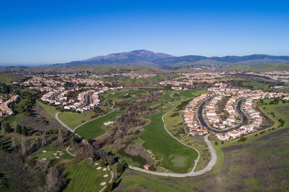 An aerial view of a residential area with winding roads, green fields, and a mountain range in the background.