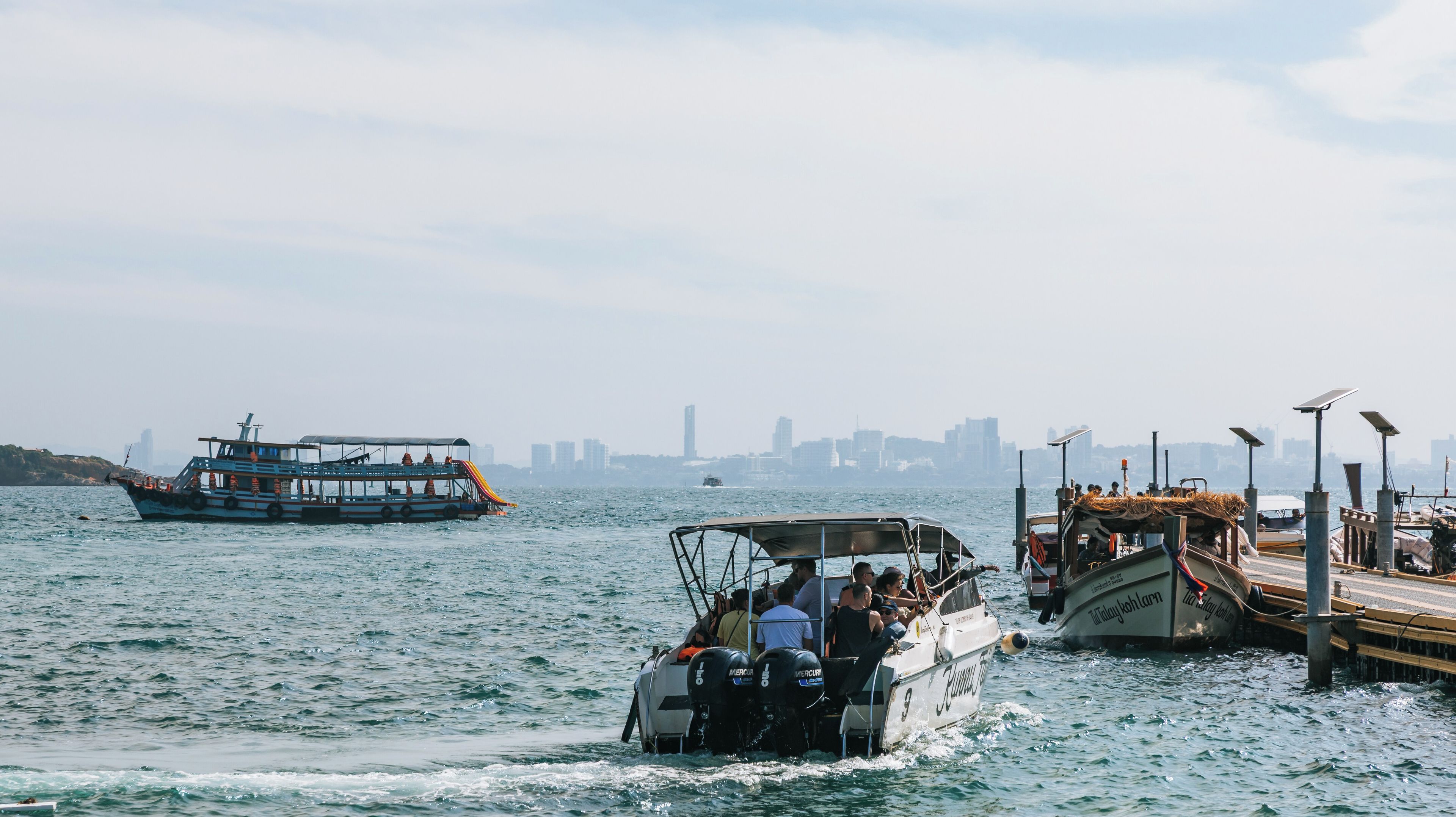 Um ferryboat com passageiros, um barco atracado com painéis solares e a silhueta de uma cidade ao fundo.