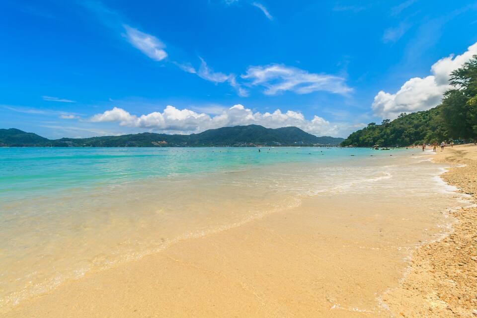 A beach with clear turquoise water, sandy shore, and green hills in the background.