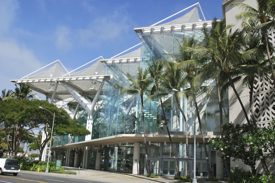 A modern glass building with a geometric design, palm trees, and a clear blue sky.