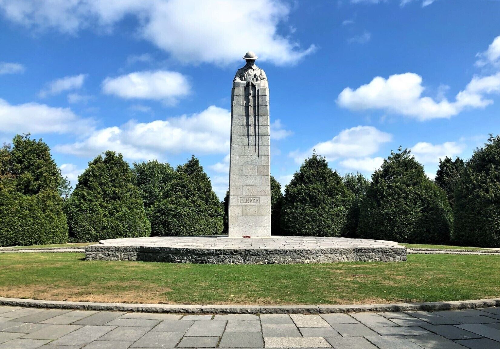 A tall monument with a plaque reading "CANADA" in a park with trees and a clear sky.