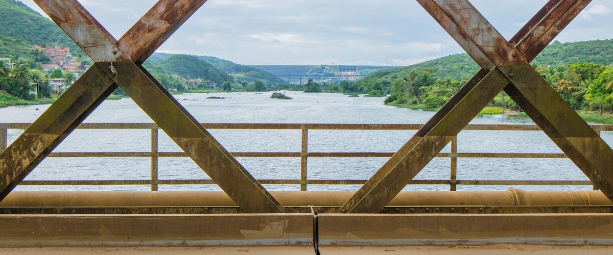 A rusted metal bridge with a river and green hills in the background.