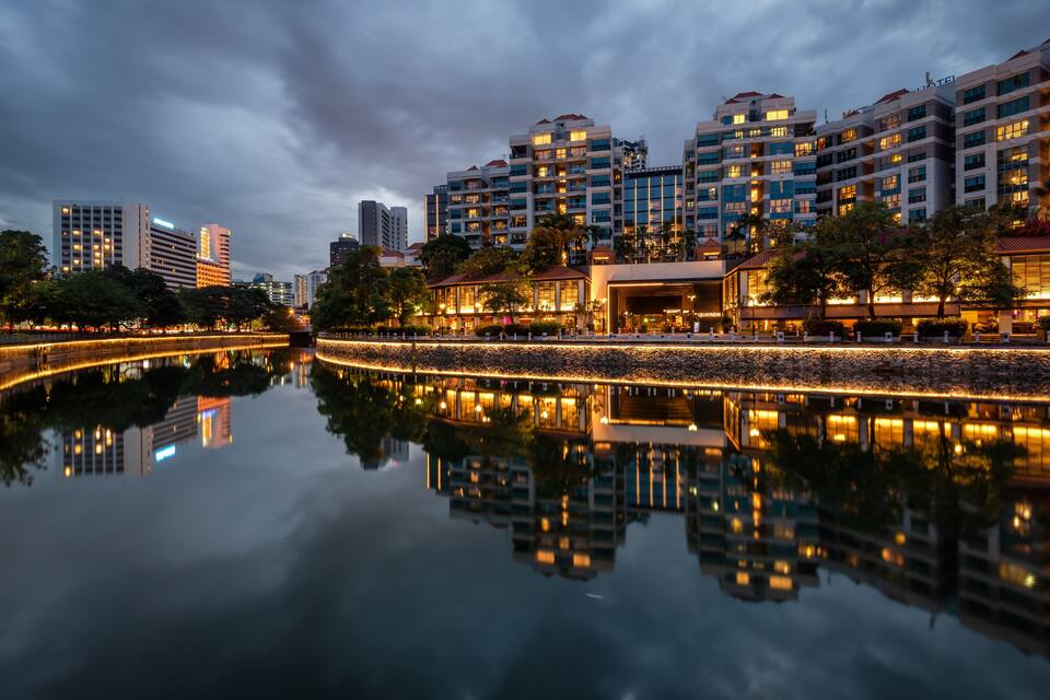 A cityscape at dusk with illuminated buildings reflecting on a calm river.