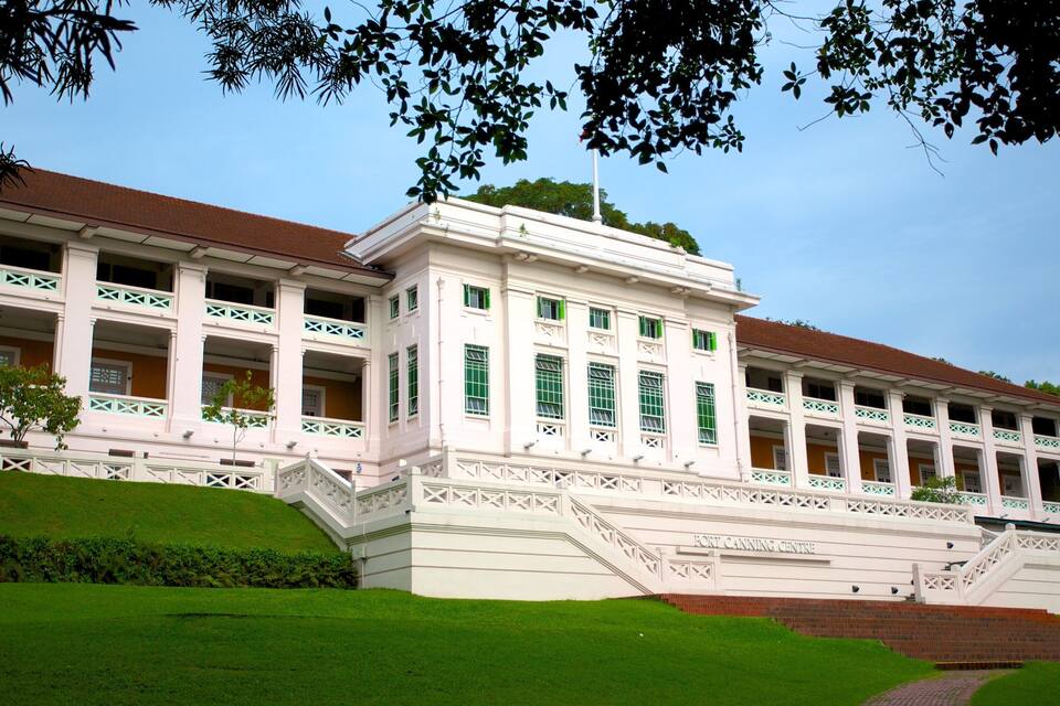 A large, white colonial-style building with green window frames and a red roof, surrounded by well-maintained green lawns and trees.