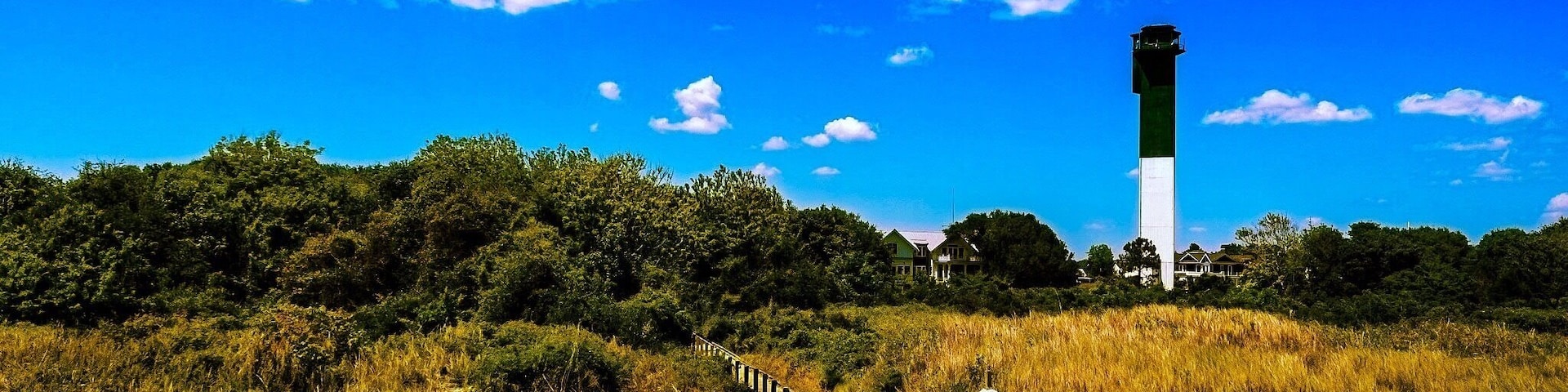 Un paseo marítimo de madera que conduce a un faro, rodeado de campos cubiertos de hierba y árboles bajo un cielo azul despejado.