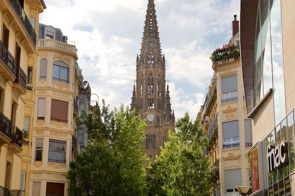 A cathedral with a tall spire, surrounded by buildings with balconies and trees.