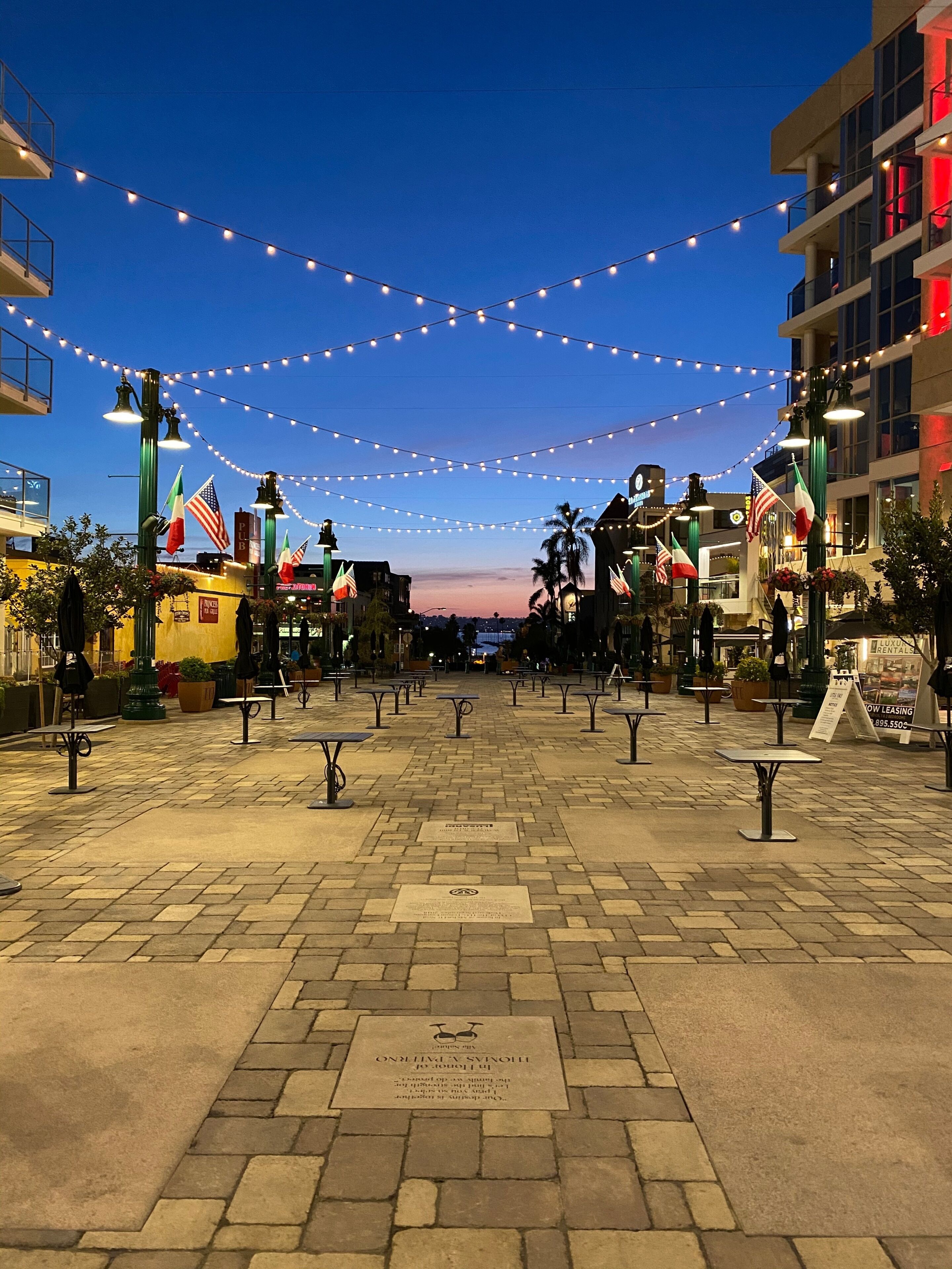 Uma praça pavimentada com luzes de Natal, bandeiras e mesas ao ar livre.