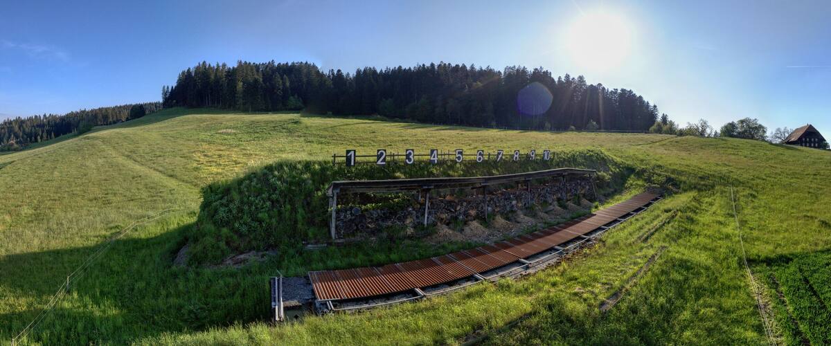 A scenic railway track winding through a lush green field with a forested hill in the background.