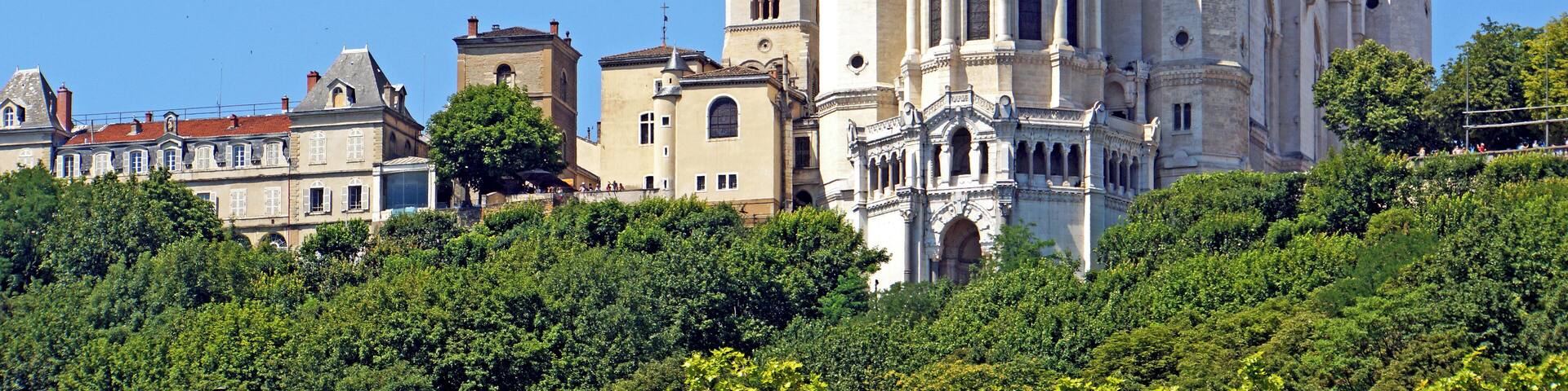 A historic cathedral with multiple spires and crosses, surrounded by lush greenery and other buildings.