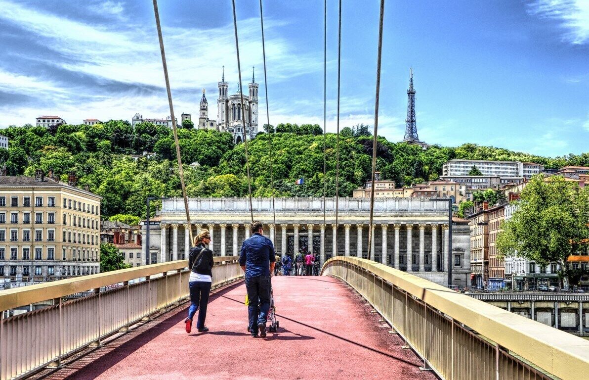 A pedestrian bridge with people walking, overlooking a city with a prominent cathedral and the Eiffel Tower in the background.