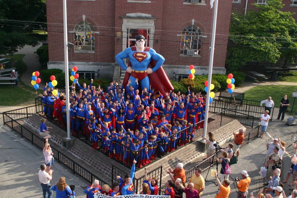 A group of people in Superman costumes gathered around a large statue in front of a brick building.