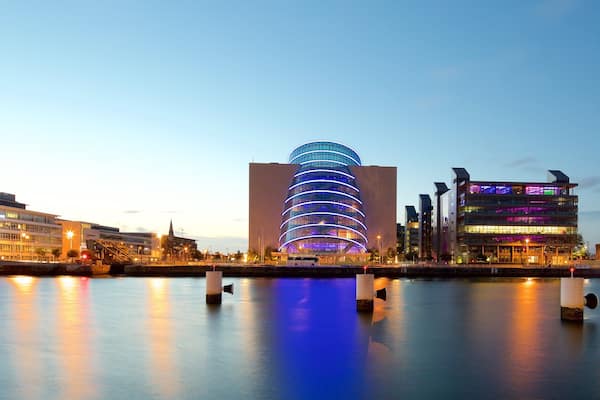 A waterfront cityscape with modern buildings, including a curved glass structure and a multi-story building with colorful lighting.