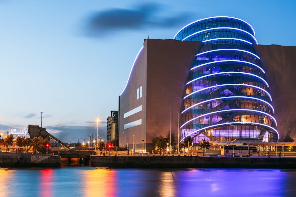 Un edifici modern i corbat amb línies blaves il·luminades, un passeig marítim i un pont al fons.