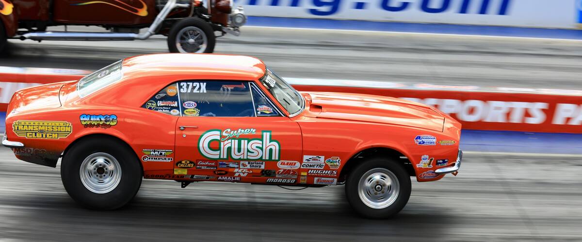 A vintage orange race car with various sponsor decals, driving on a track with a blurred background.