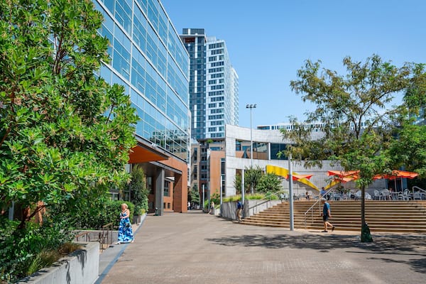 A modern urban plaza with glass buildings, outdoor seating, and green trees.