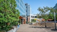 Una plaza urbana moderna con edificios de vidrio, mesas al aire libre y árboles verdes.