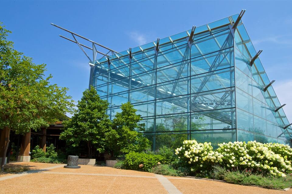 Un bâtiment moderne en verre, doté d’une structure métallique, entouré d’un jardin aménagé où poussent diverses plantes et fleurs.