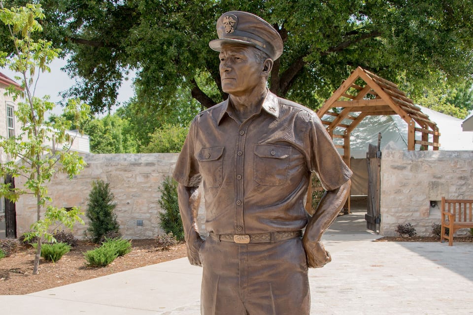 A bronze statue of a man in a hat and uniform standing on a paved area with trees and a stone wall in the background.