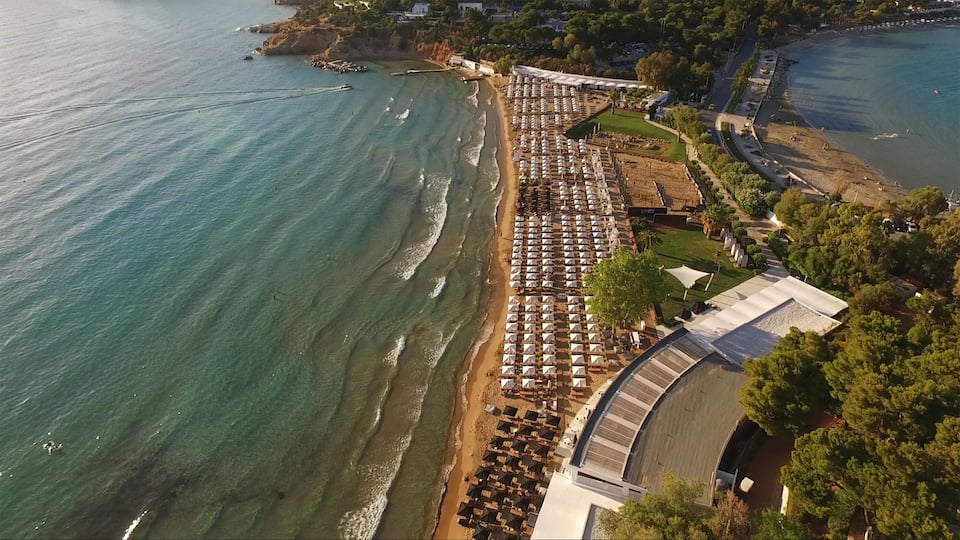 An aerial view of a coastal area with a beach lined with umbrellas and a large building with a curved roof.