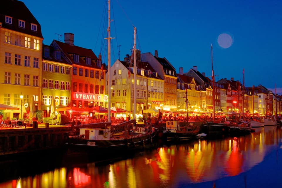 A waterfront with buildings, boats, and a moon in the sky.