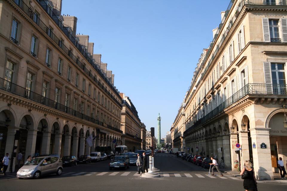 A street lined with historic buildings, a tower in the distance, and parked cars.