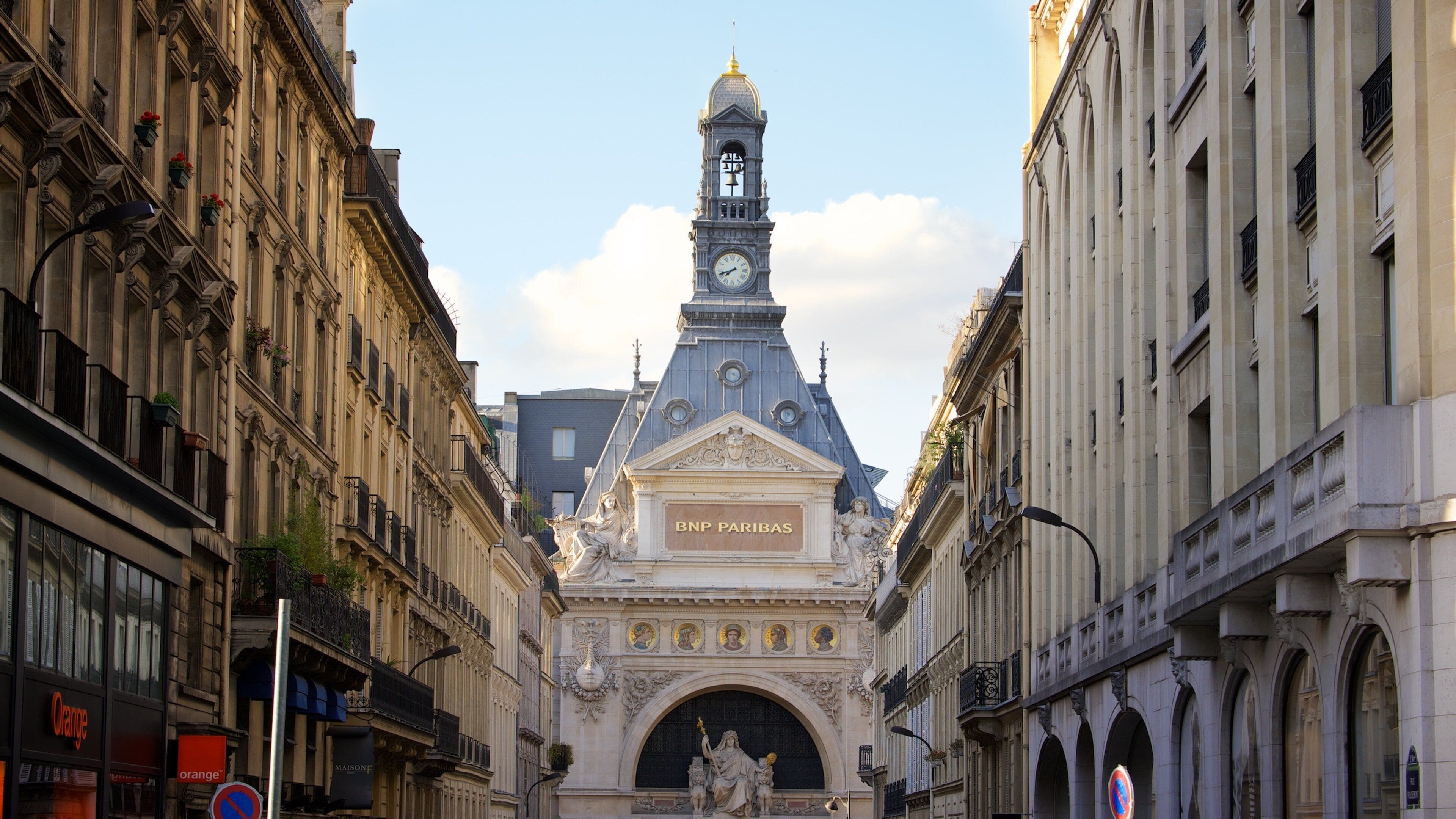 Calle con edificios ornamentados, una torre del reloj y una estatua de una mujer sosteniendo a un niño.