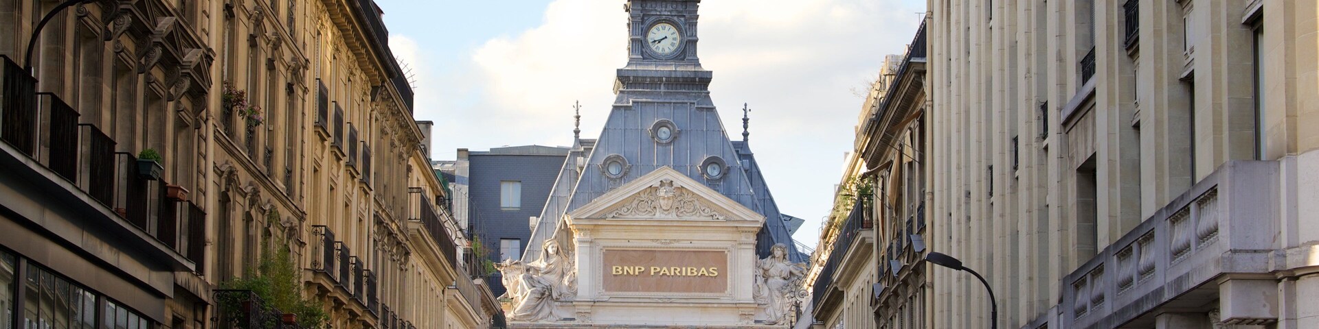 Calle con edificios ornamentados, una torre del reloj y una estatua de una mujer sosteniendo a un niño.