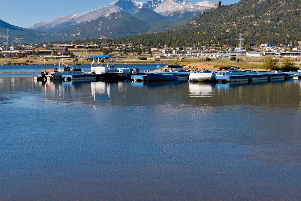 Un llac tranquil amb diverses embarcacions amarrades, una serralada de muntanyes al fons i un petit poble amb cases i edificis.