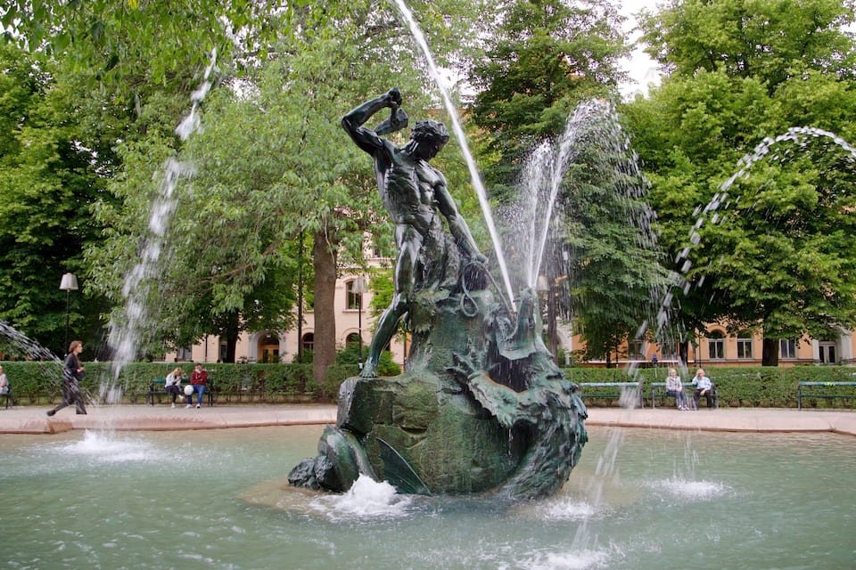 Una estatua de un hombre sosteniendo una espada en un parque con fuentes y árboles.