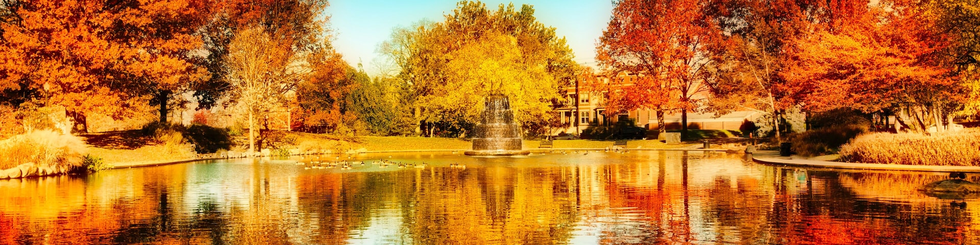A serene lake with autumn foliage, a fountain, and a clear blue sky.