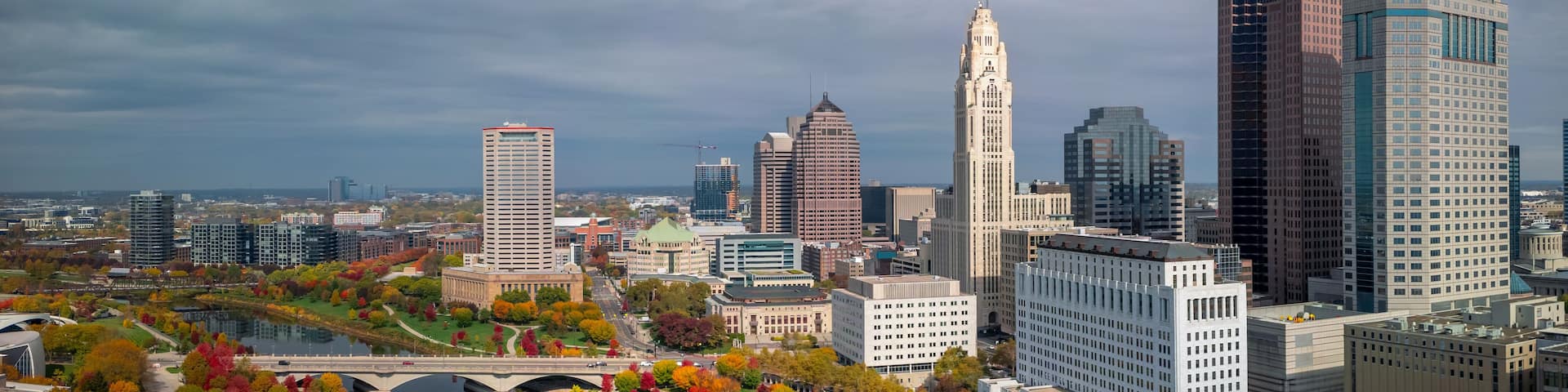 A city skyline with a prominent clock tower, modern skyscrapers, and a river with autumn foliage.