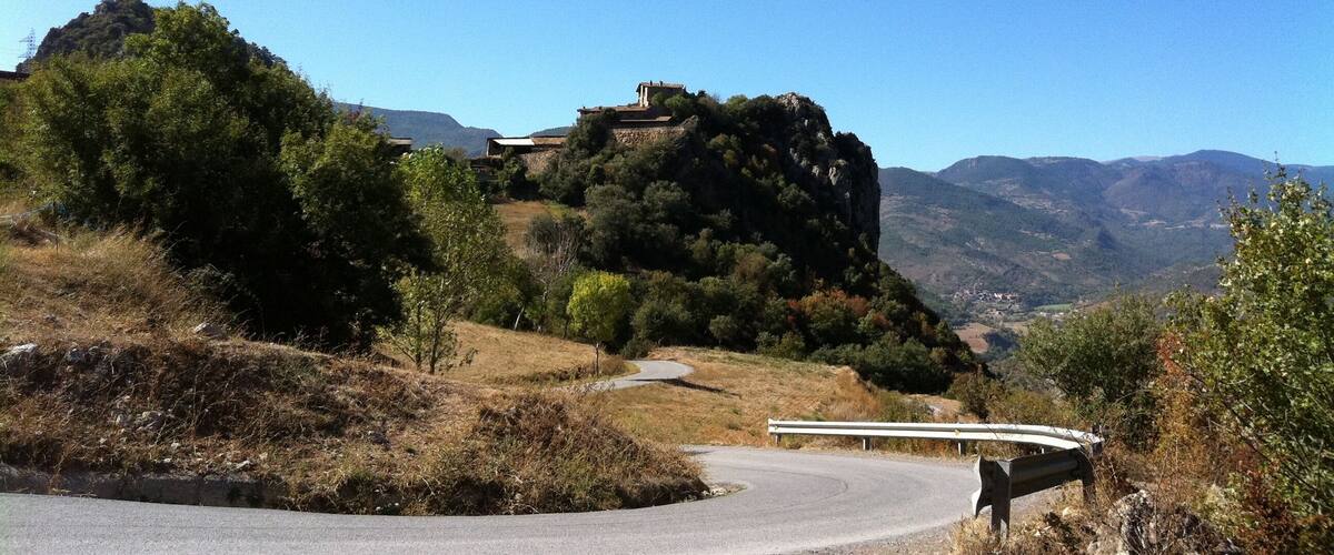 A winding road with a guardrail, a rocky hill with sparse vegetation, and distant mountains under a clear blue sky.