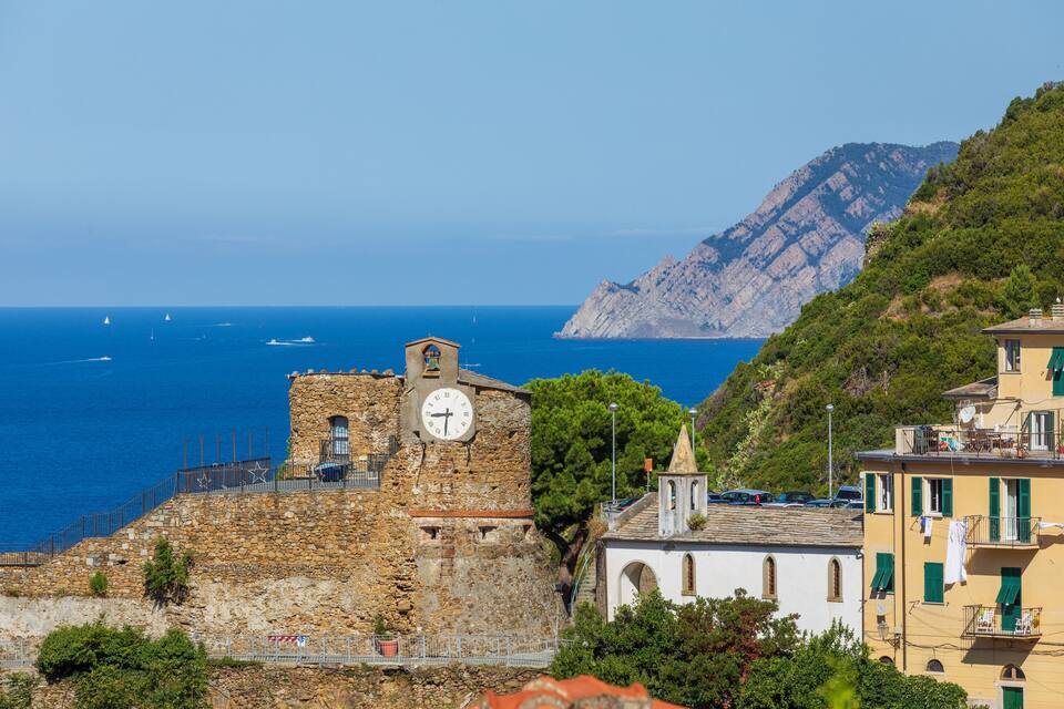 A coastal town with a historic stone building, a clock tower, and a mountainous backdrop.