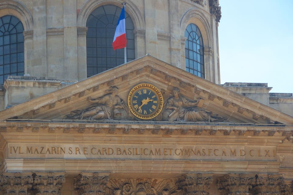 Fachada d'edifici amb un rellotge gran, la bandera francesa i treballs decoratius ornamentals.