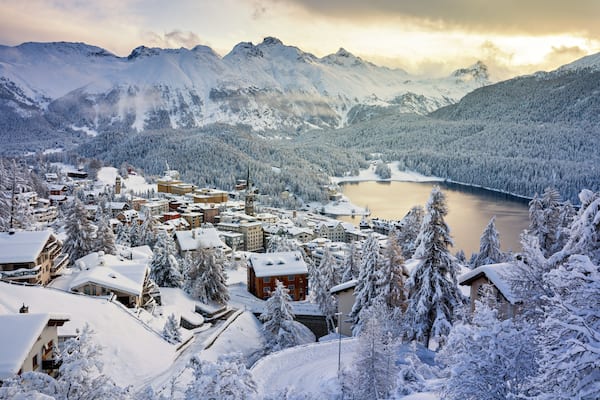 A snow-covered mountain town with a lake and forested mountains in the background.