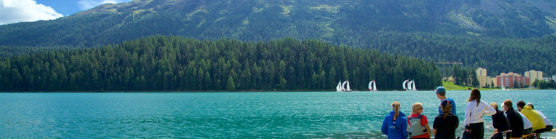 A lake with sailboats, a forested mountain, and people on the shore.