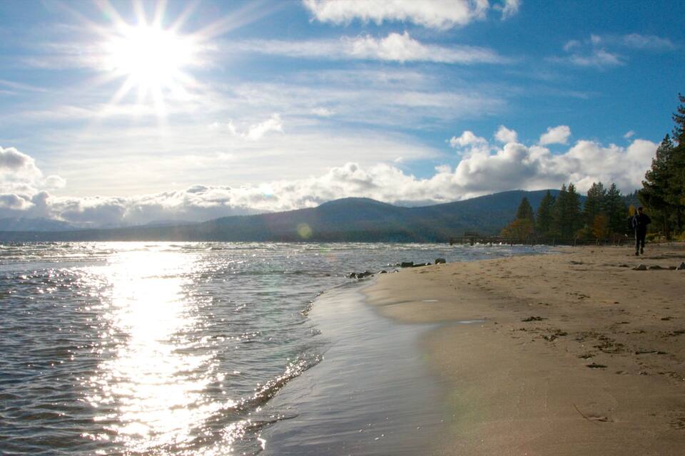 A beach with a clear sky, sunlight reflecting on the water, and mountains in the background.