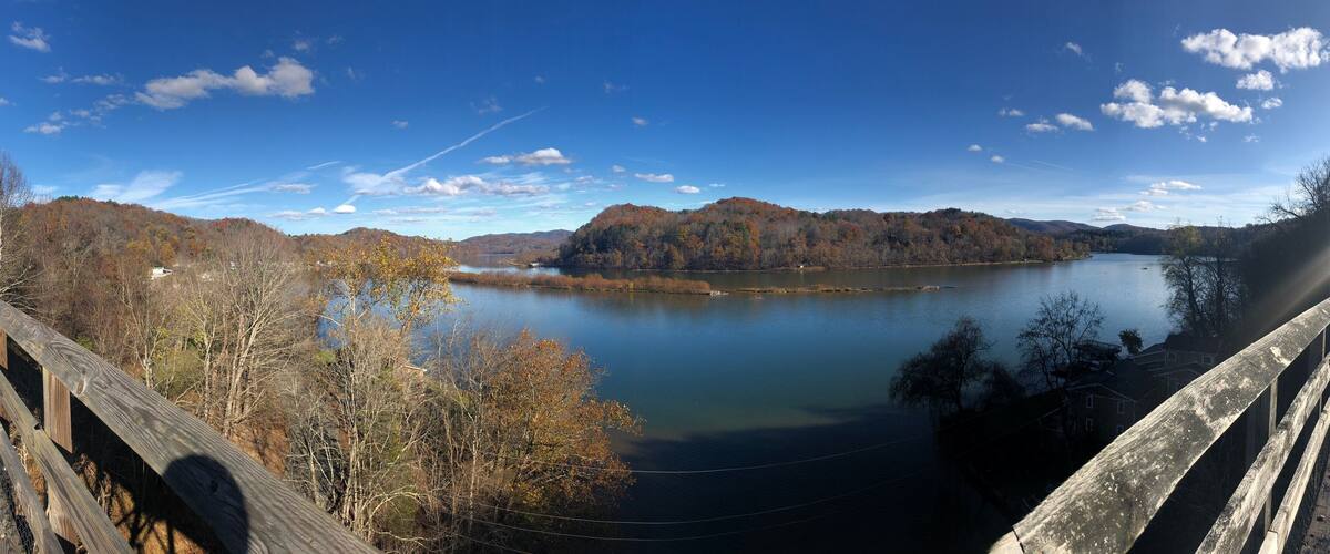 A wooden bridge over a calm lake surrounded by autumn-colored trees and hills.