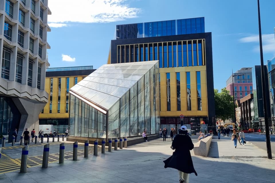 Moderni kaupunkiaukio, jossa on nykyaikaisia rakennuksia, lasirakenteita ja jalankulkijoita.