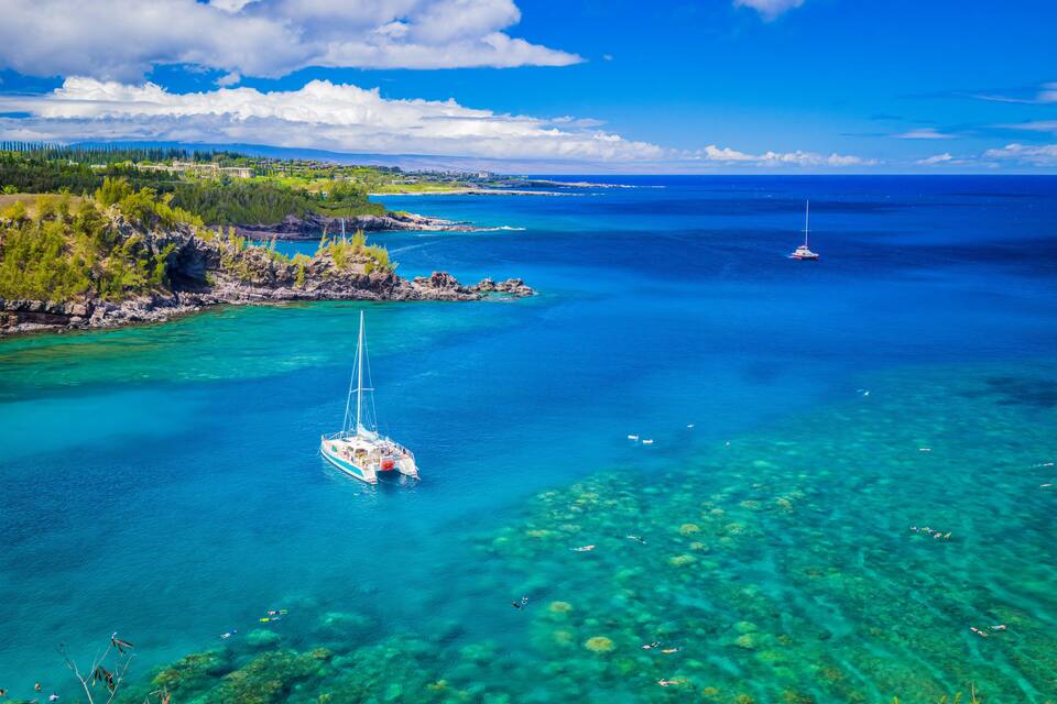 Une mer d’un bleu profond, avec un voilier, une côte rocheuse et une terre lointaine à l’horizon.
