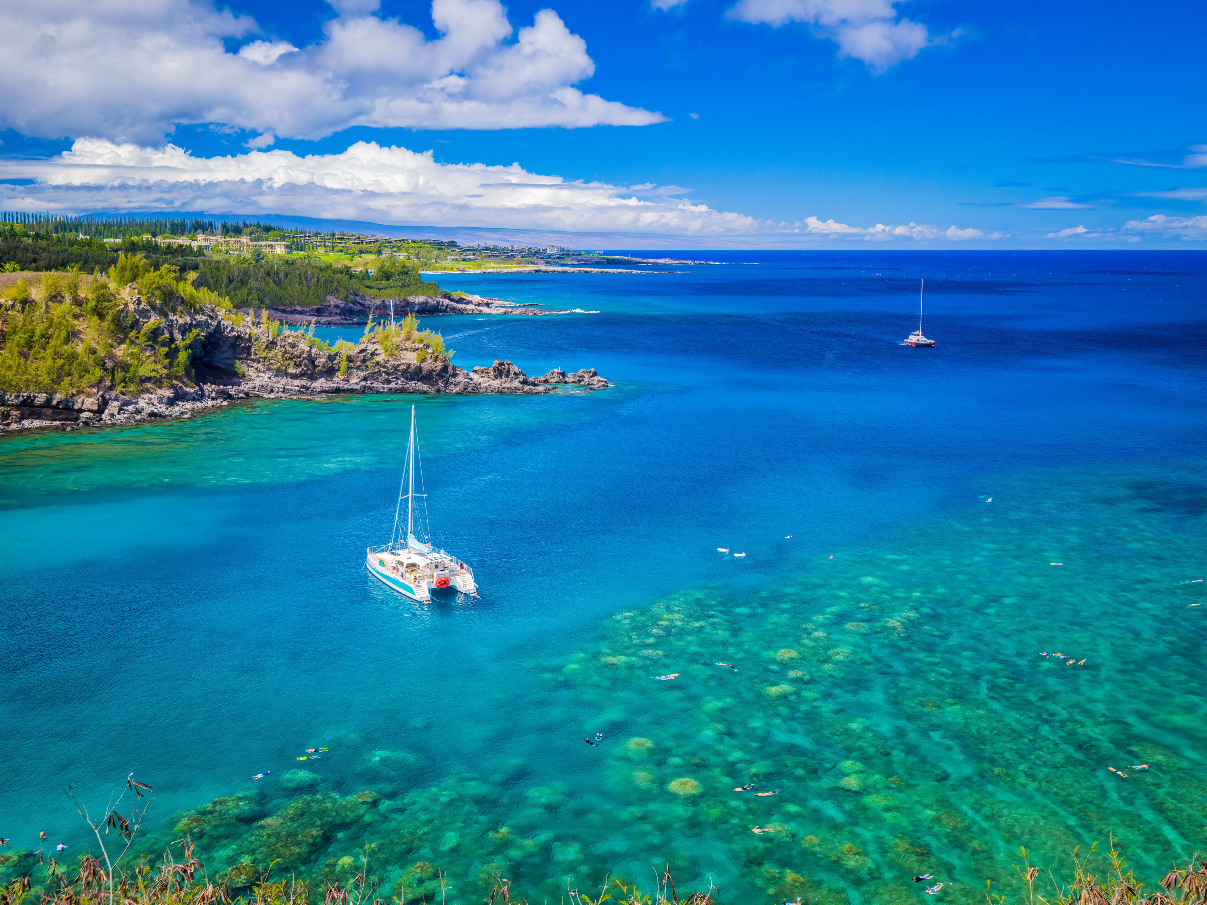 A clear blue sea with a sailboat, rocky coastline, and distant land.