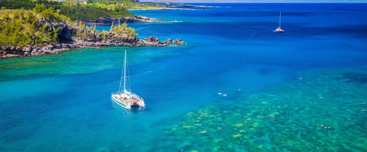 A clear blue sea with a sailboat, rocky coastline, and distant land.