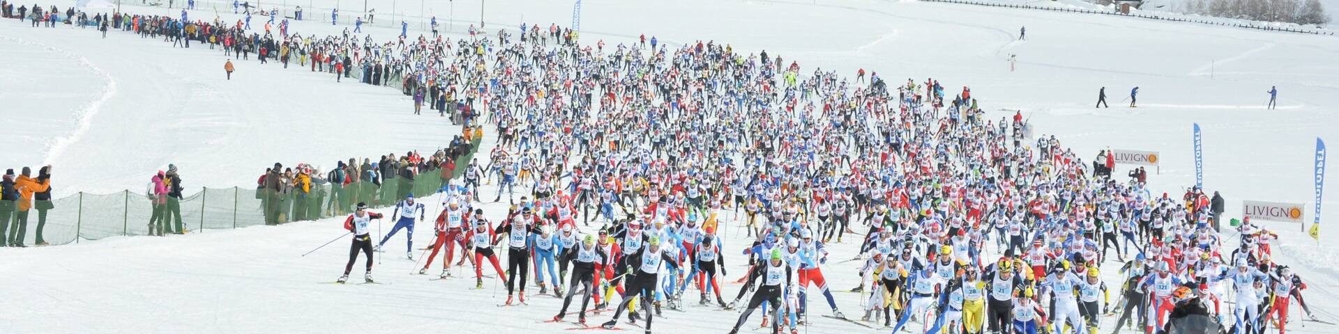 A large group of skiers on a snowy slope with buildings in the background.