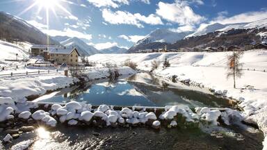 A snow-covered landscape with a clear stream, mountains in the background, and a wooden house by the stream.