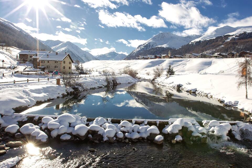 Lumipeitteinen maisema, jossa virtaa kirkas puro, taustalla kohoavat vuoret ja puron varrella sijaitsee puinen talo.