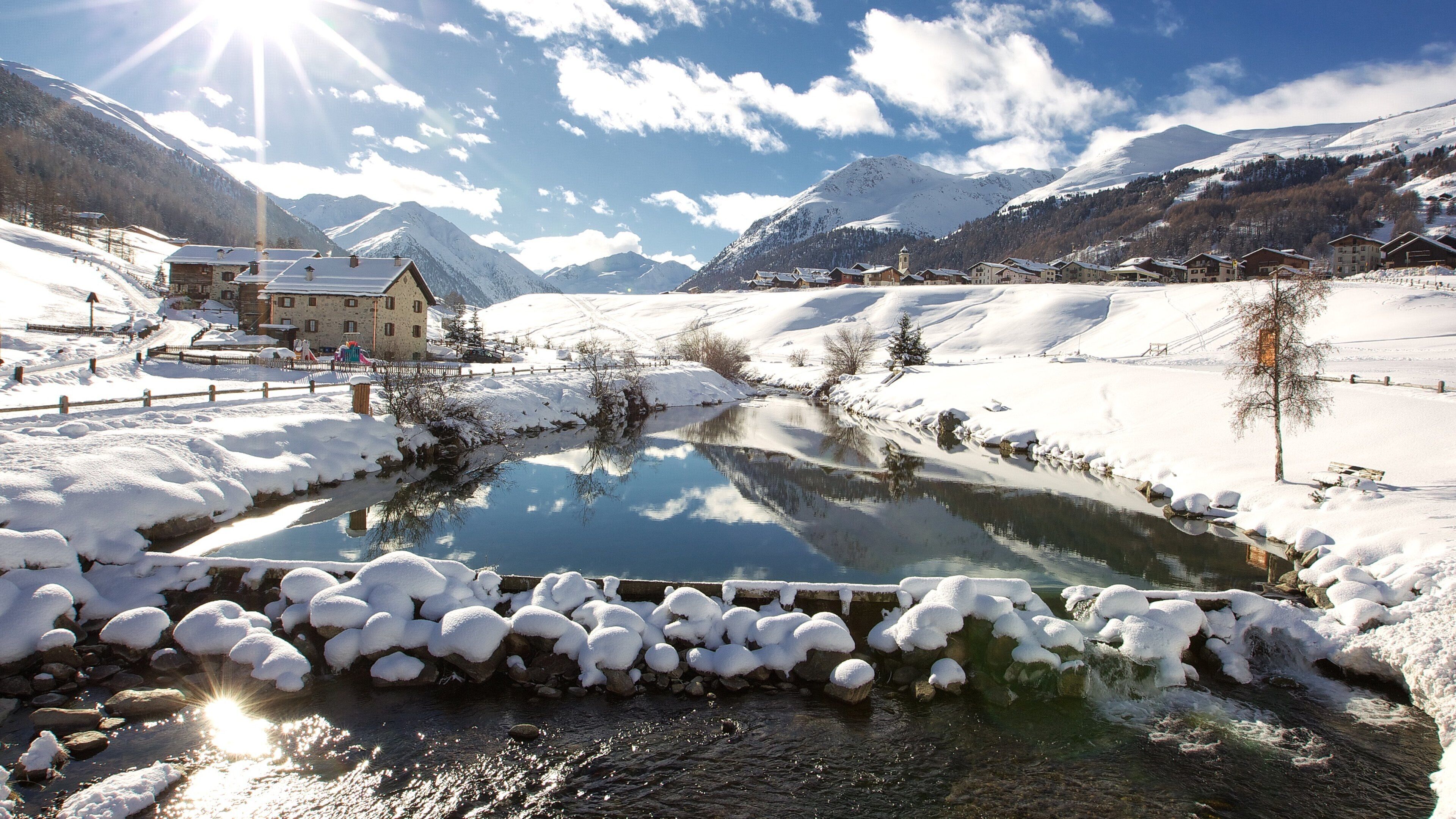 A snow-covered landscape with a clear stream, mountains in the background, and a wooden house by the stream.