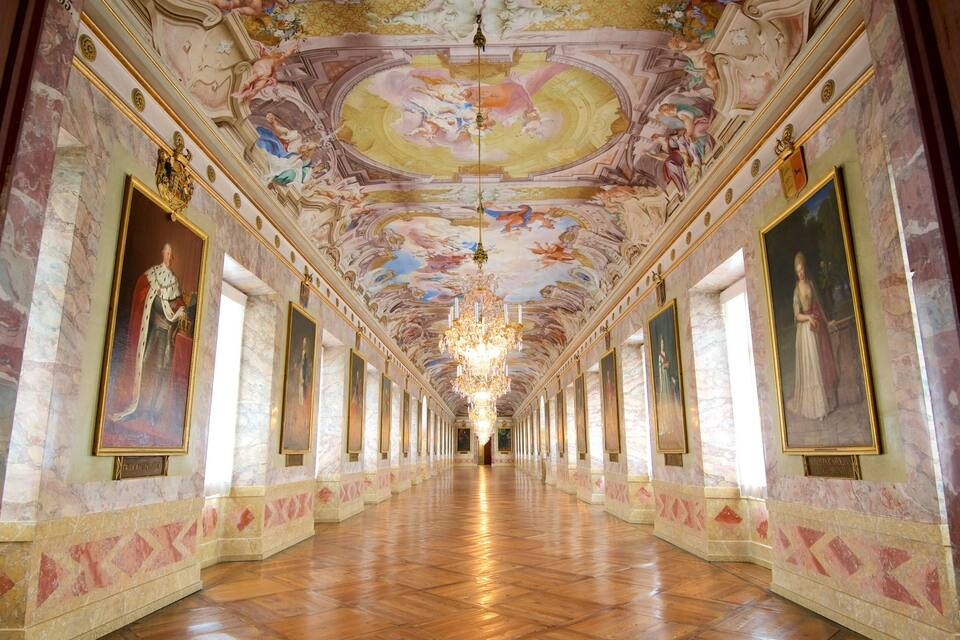 A grand hallway with ornate ceiling frescoes, a large chandelier, and classical paintings on the walls.