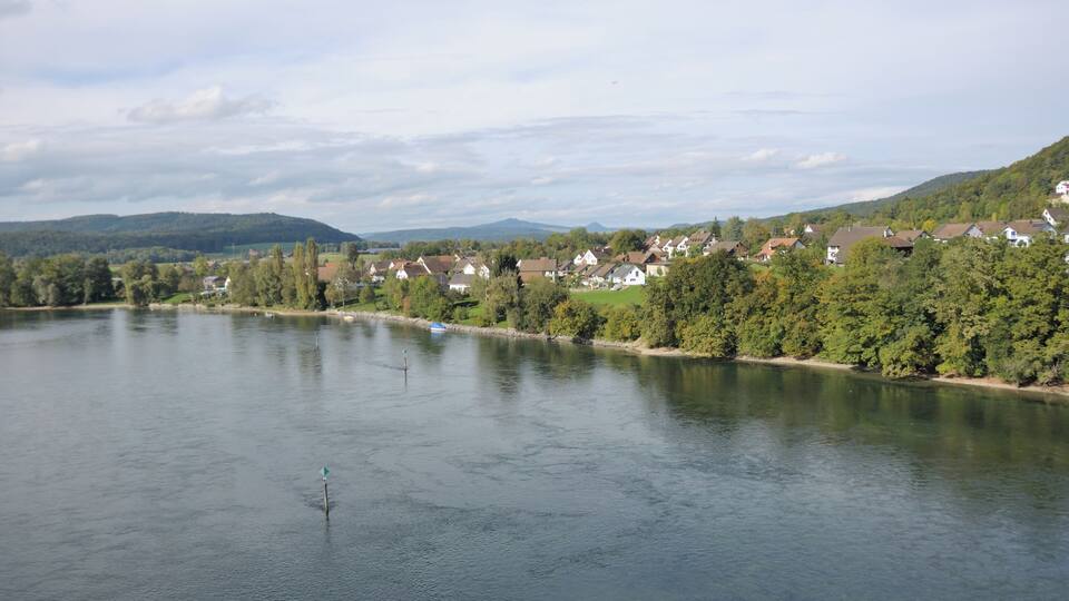 A calm river with houses along the banks, surrounded by green hills and trees.